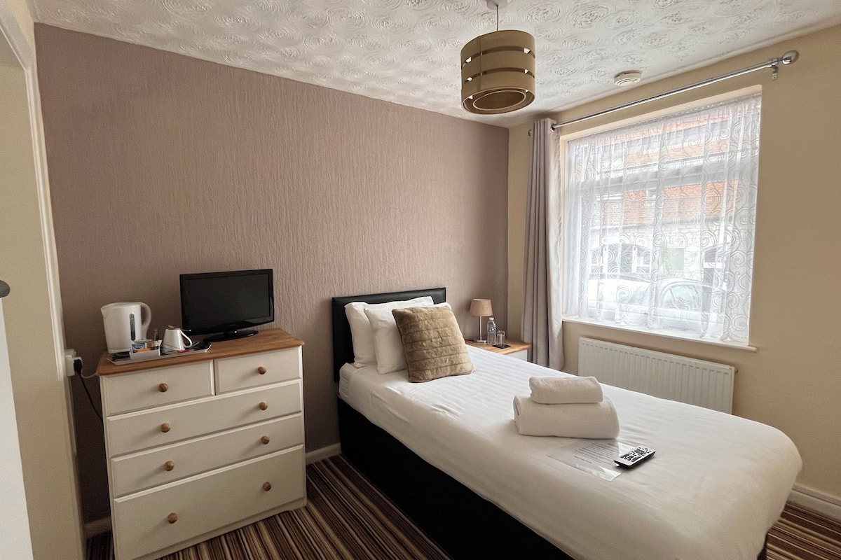 A hotel room with a bed, a TV on a wooden dresser, a window with curtains, and a radiator.