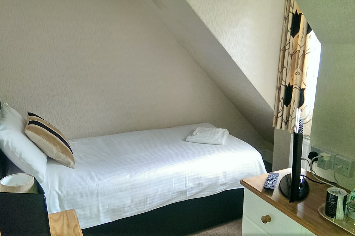 A small, neatly made bed in a corner room with a sloped ceiling. There is a bedside table with a remote control, a lamp, and some toiletries.