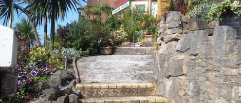 Un escalier menant à l’entrée d’un hôtel, avec un mur de pierre d’un côté et une variété de plantes et de fleurs de l’autre.