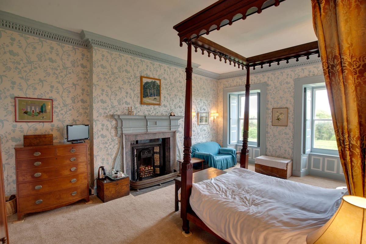 A bedroom with a four-poster bed, a fireplace, a dresser, and a window with curtains.