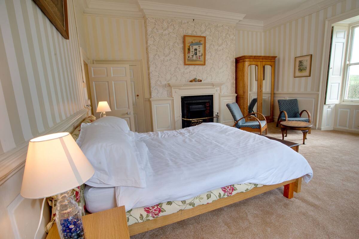 A neatly made bed with white linens, a bedside lamp, a floral-patterned pillow, and a small table with a jar of decorative items.