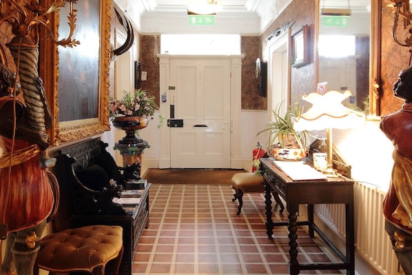 A hallway with a checkered floor, a white door at the end, and a table with a lamp and decorative items.