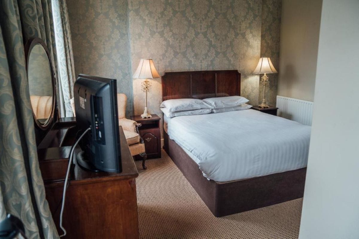 Quarto de hotel com cama, televisão, mesa de cabeceira e luminária.