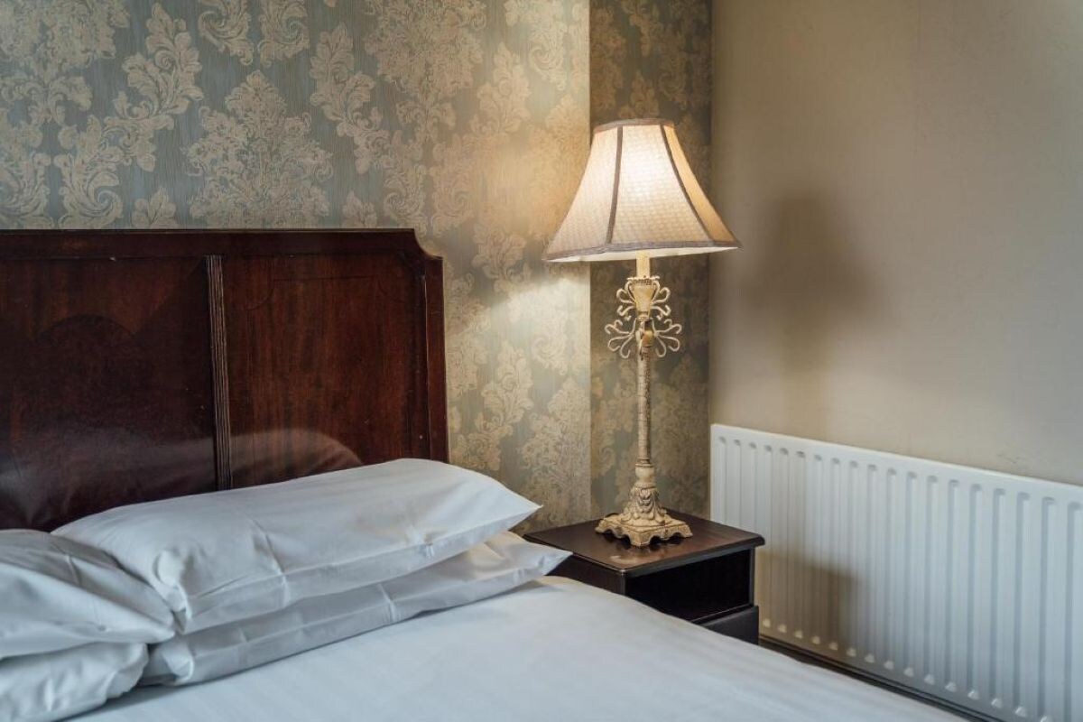 A neatly made bed with white linens, a wooden headboard, a small bedside table with a lamp, and a radiator on the wall.