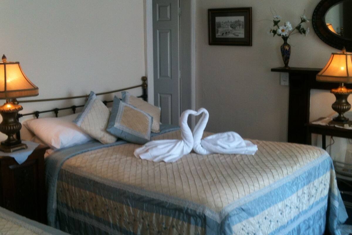 A neatly made bed with a heart-shaped towel, a bedside lamp, and a framed picture on the wall.