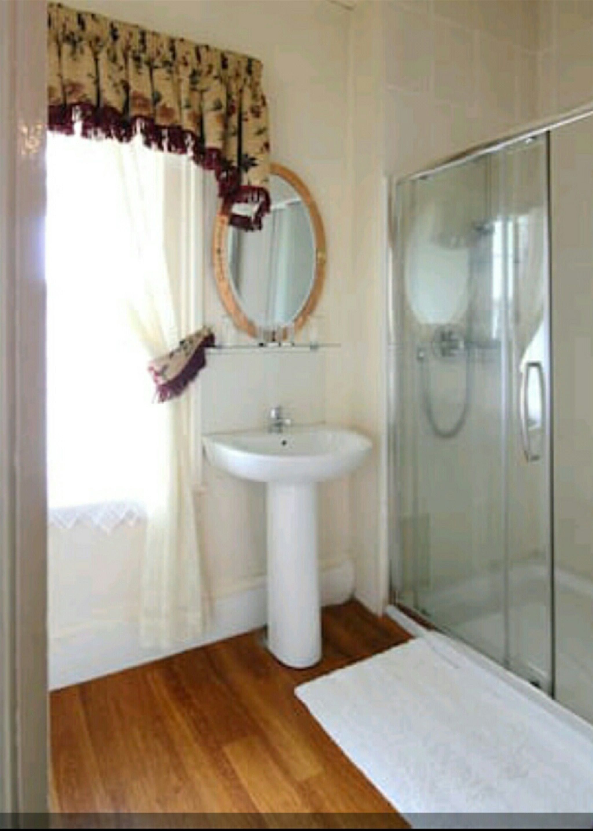 A bathroom with a pedestal sink, a glass shower enclosure, a mirror with a wooden frame, and a window with a floral curtain.