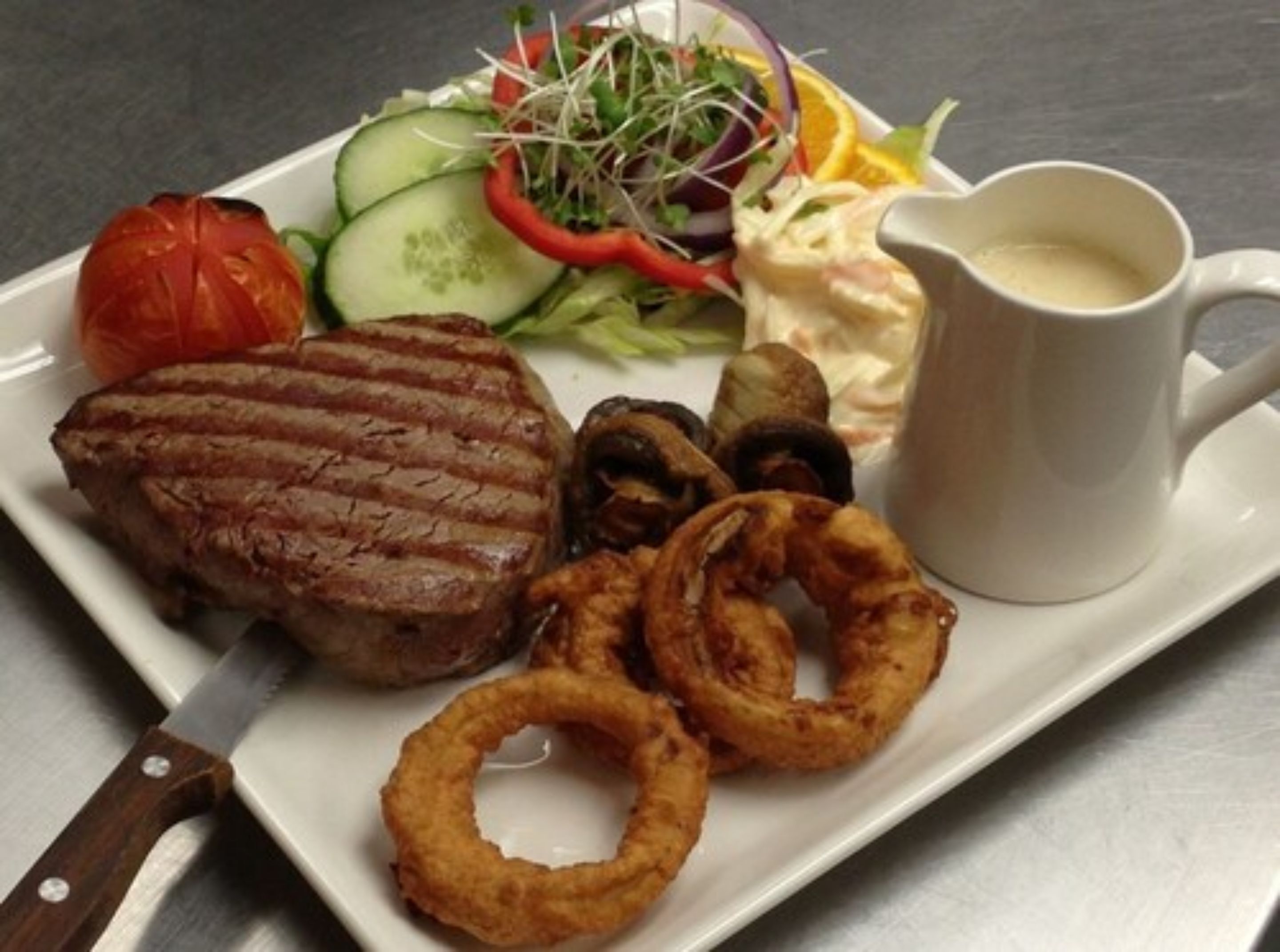 A plate with a steak, onion rings, a salad, and a cup of sauce.