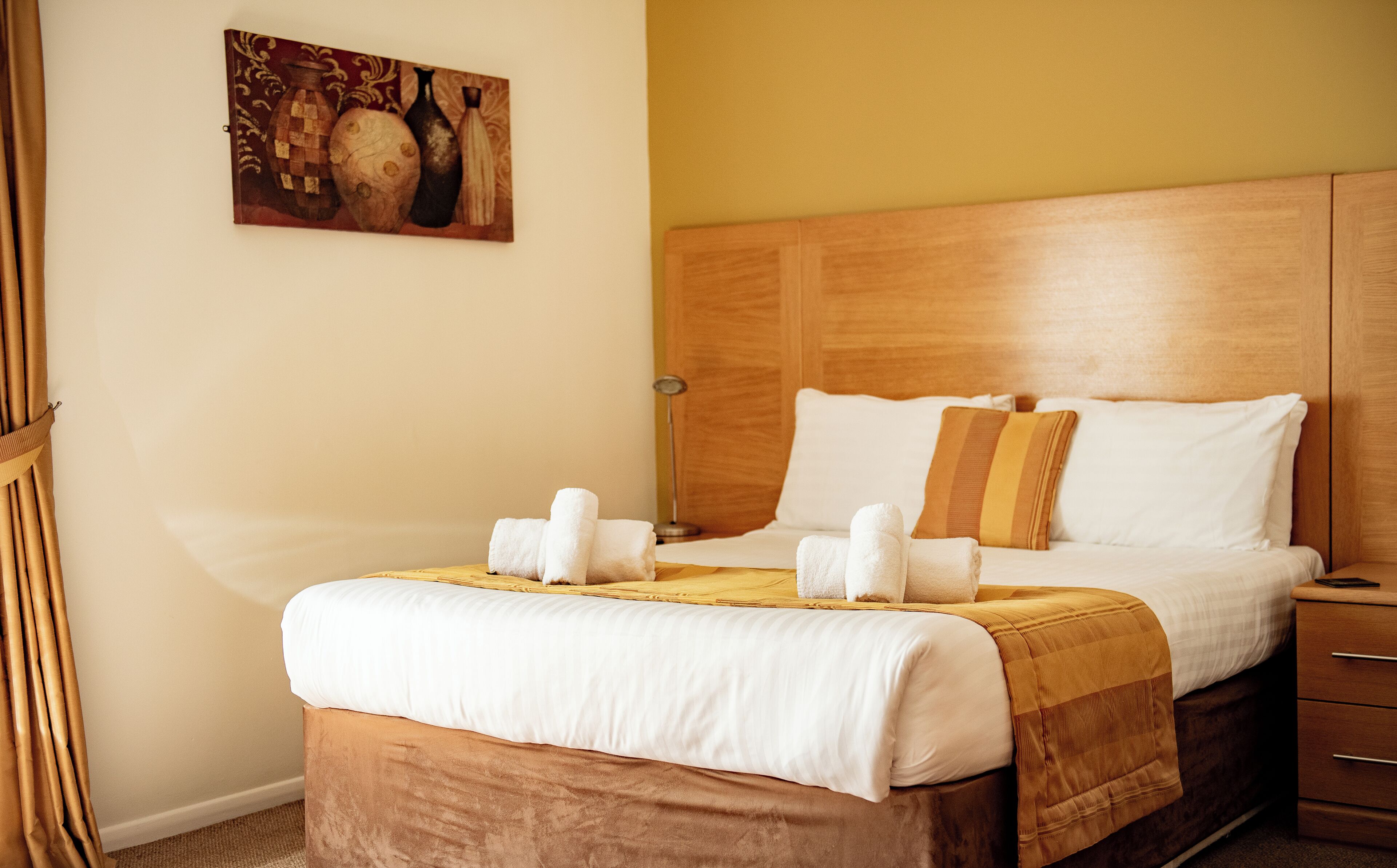 A neatly made bed with white linens and a brown headboard, a bedside table with a lamp, and a painting on the wall.