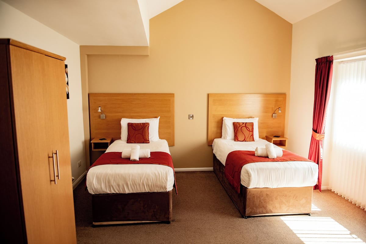 Une chambre d’hôtel avec deux lits, des têtes de lit en bois, une armoire et une fenêtre avec des rideaux.