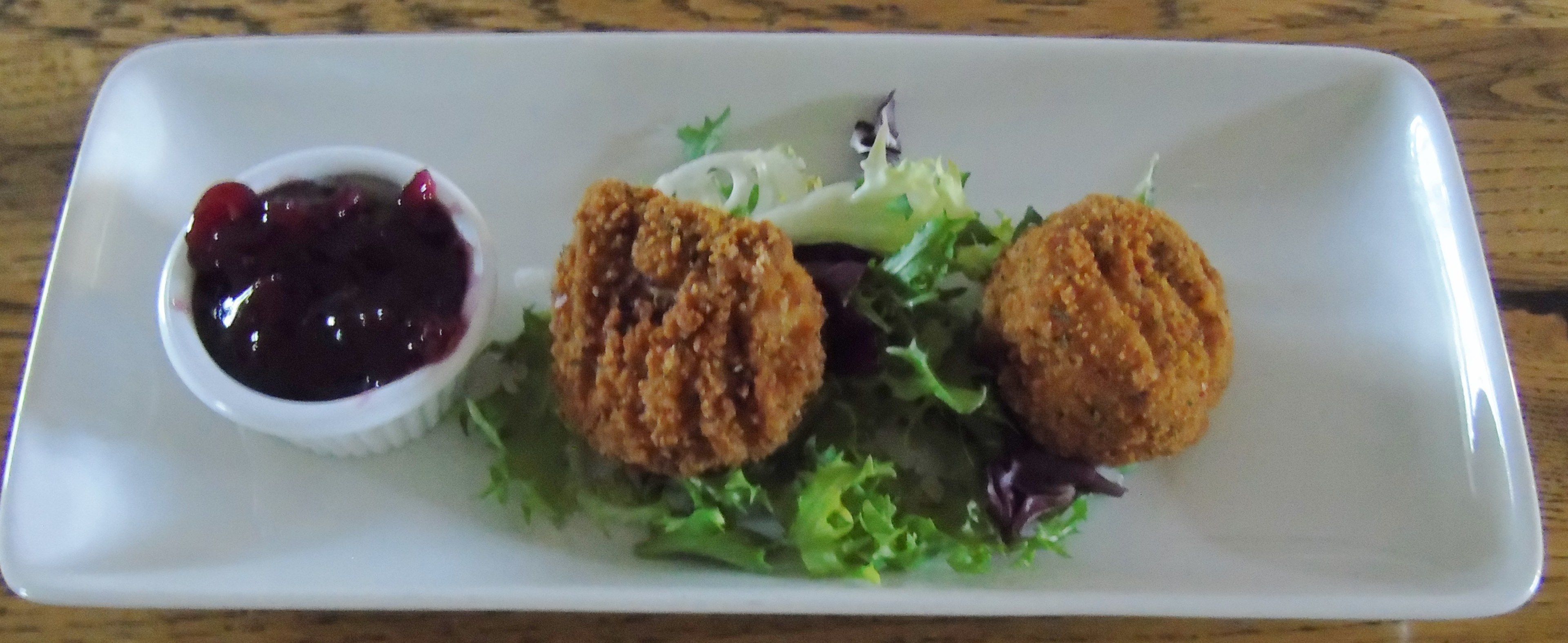 Ein rechteckiger, weißer Teller mit zwei gebratenen Speisen, einer kleinen Schale dunkellrote Sauce und einem Salat aus grünen und violetten Blättern.