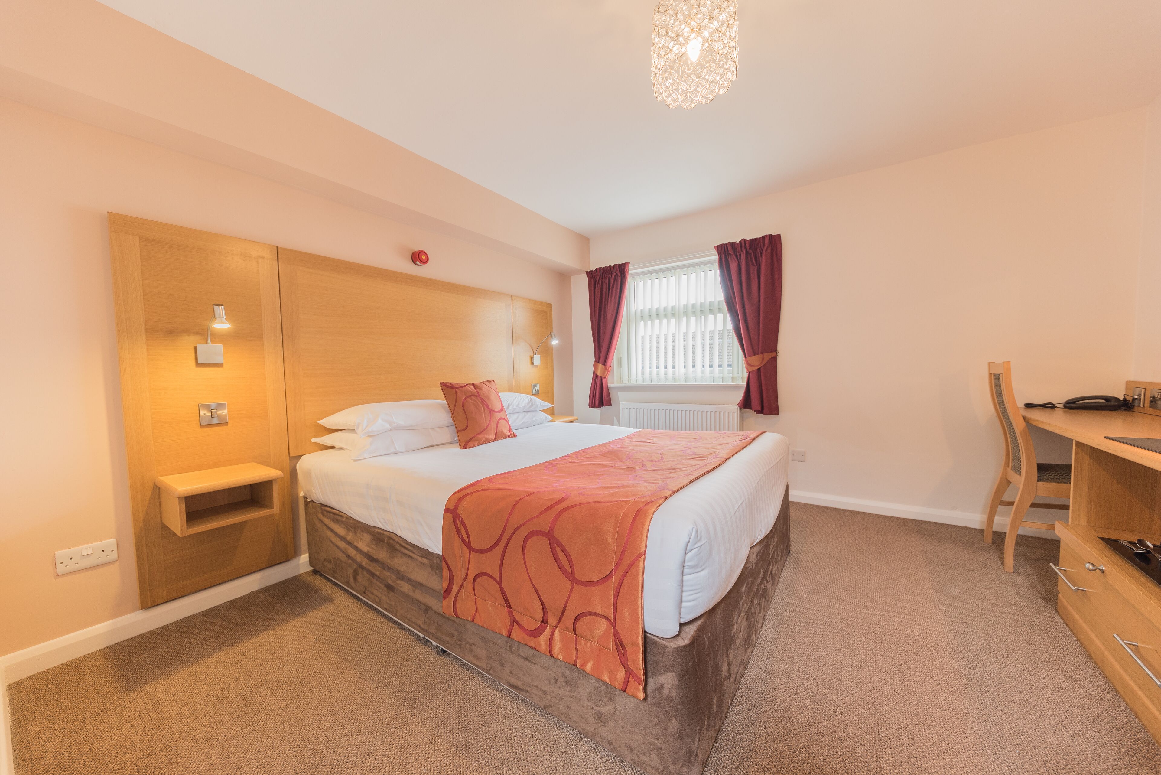 A hotel room with a bed, bedside table, lamp, and a desk with a chair.