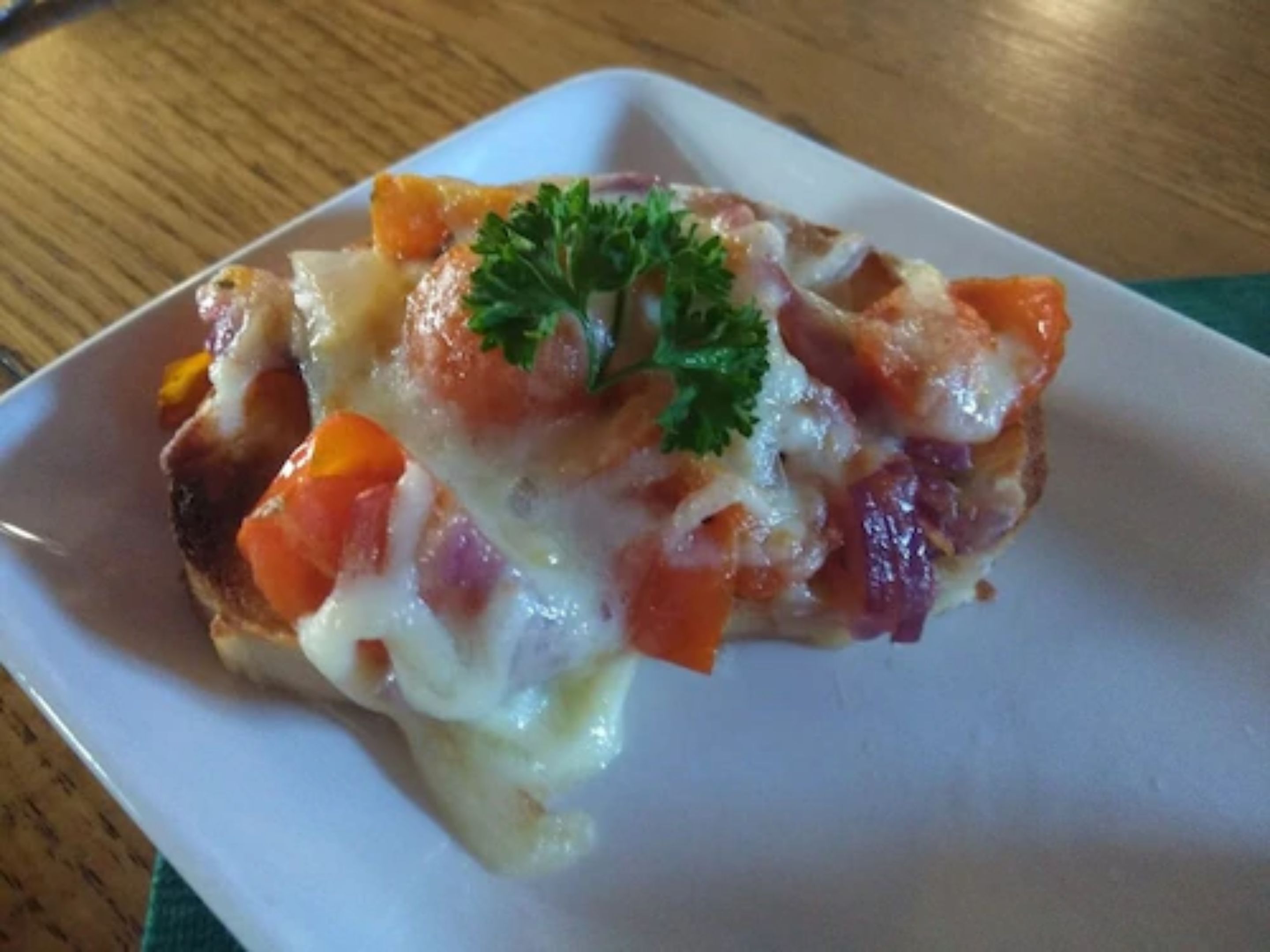 A square plate with a grilled sandwich topped with melted cheese and a garnish of parsley.