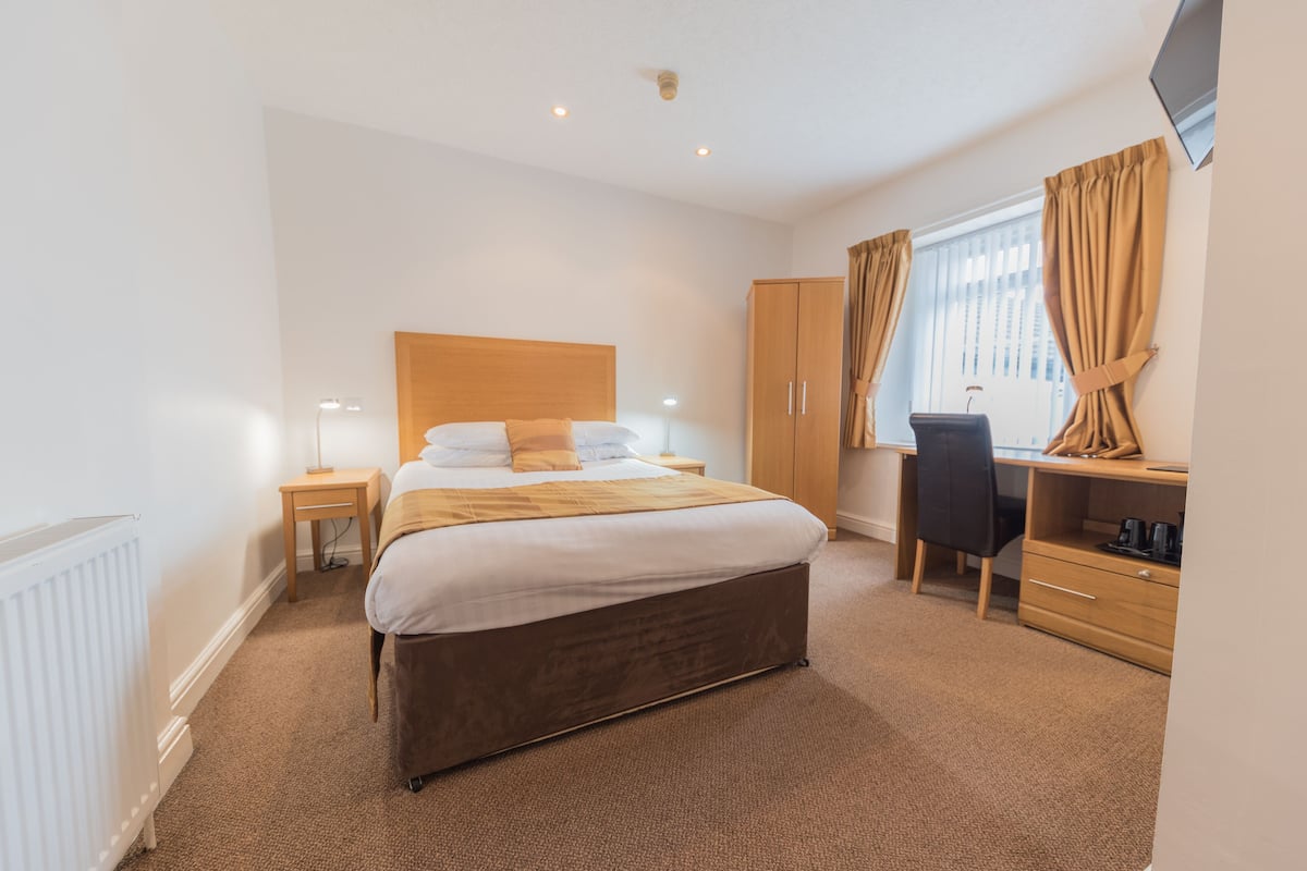 Une chambre d’hôtel avec un lit, un bureau, une chaise, une armoire et une fenêtre avec des rideaux.