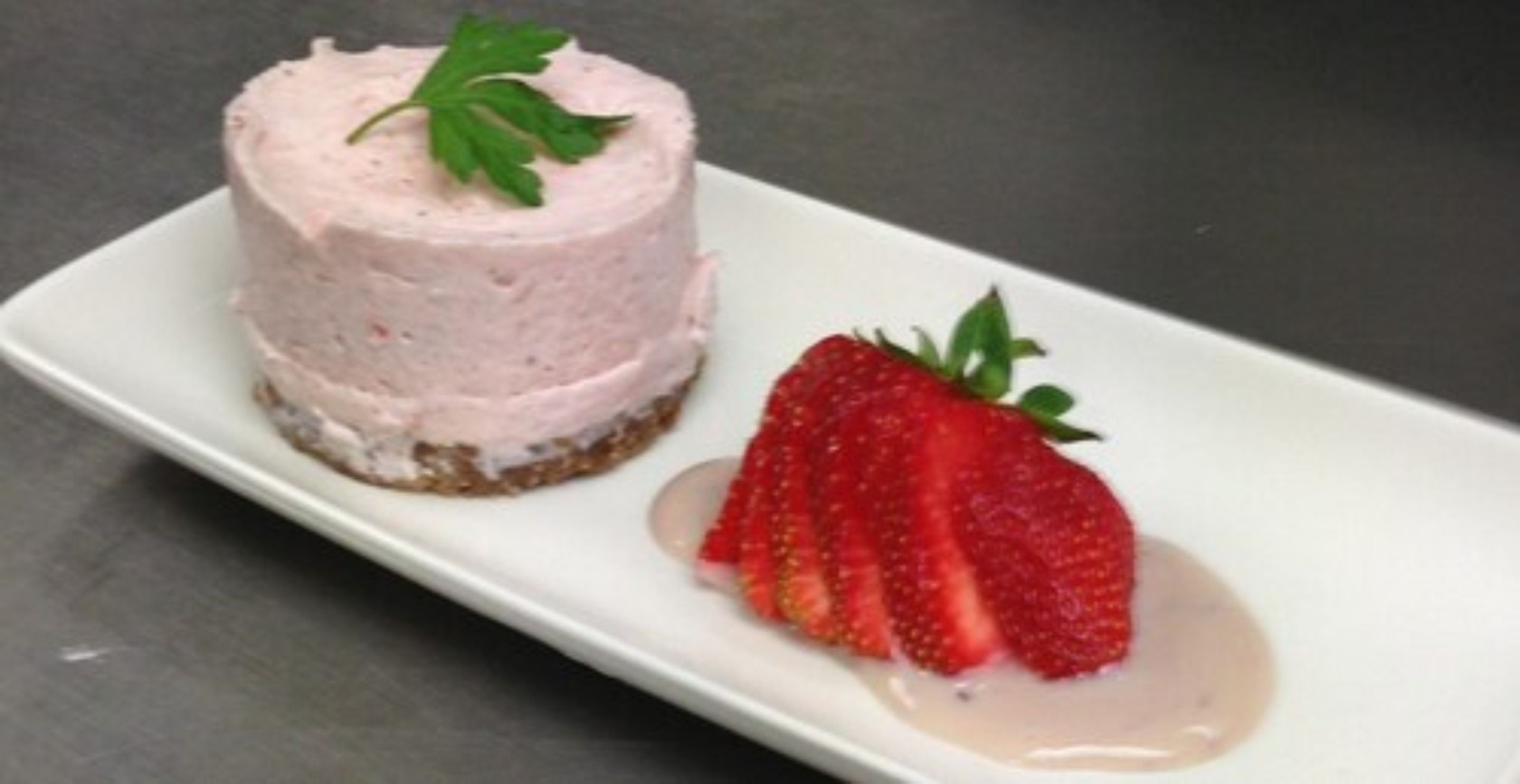 Postre de mousse rosa sobre una base, acompanyat d'una fresa i una petita cullada de crema.