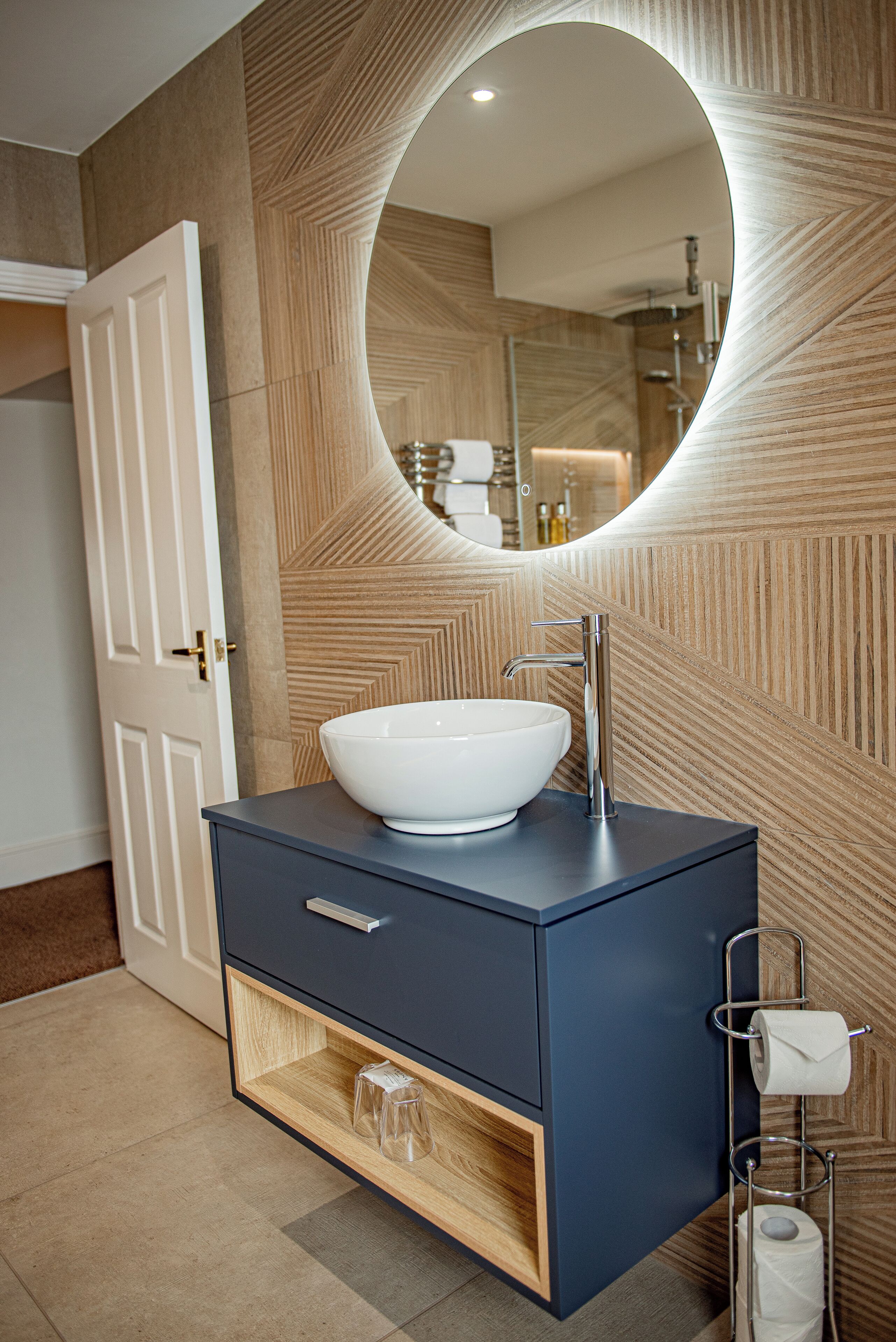 A modern bathroom with a large oval mirror, a white vessel sink, and a dark blue vanity with a wooden cabinet.
