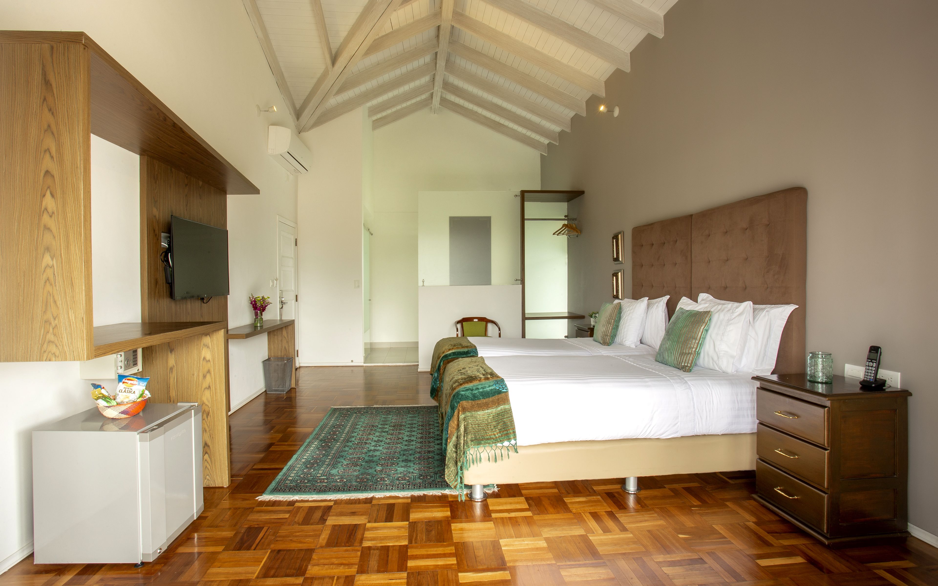 A hotel room with a wooden floor, a bed with white bedding, a wooden dresser, a small desk with a TV, and a mirror on the wall.