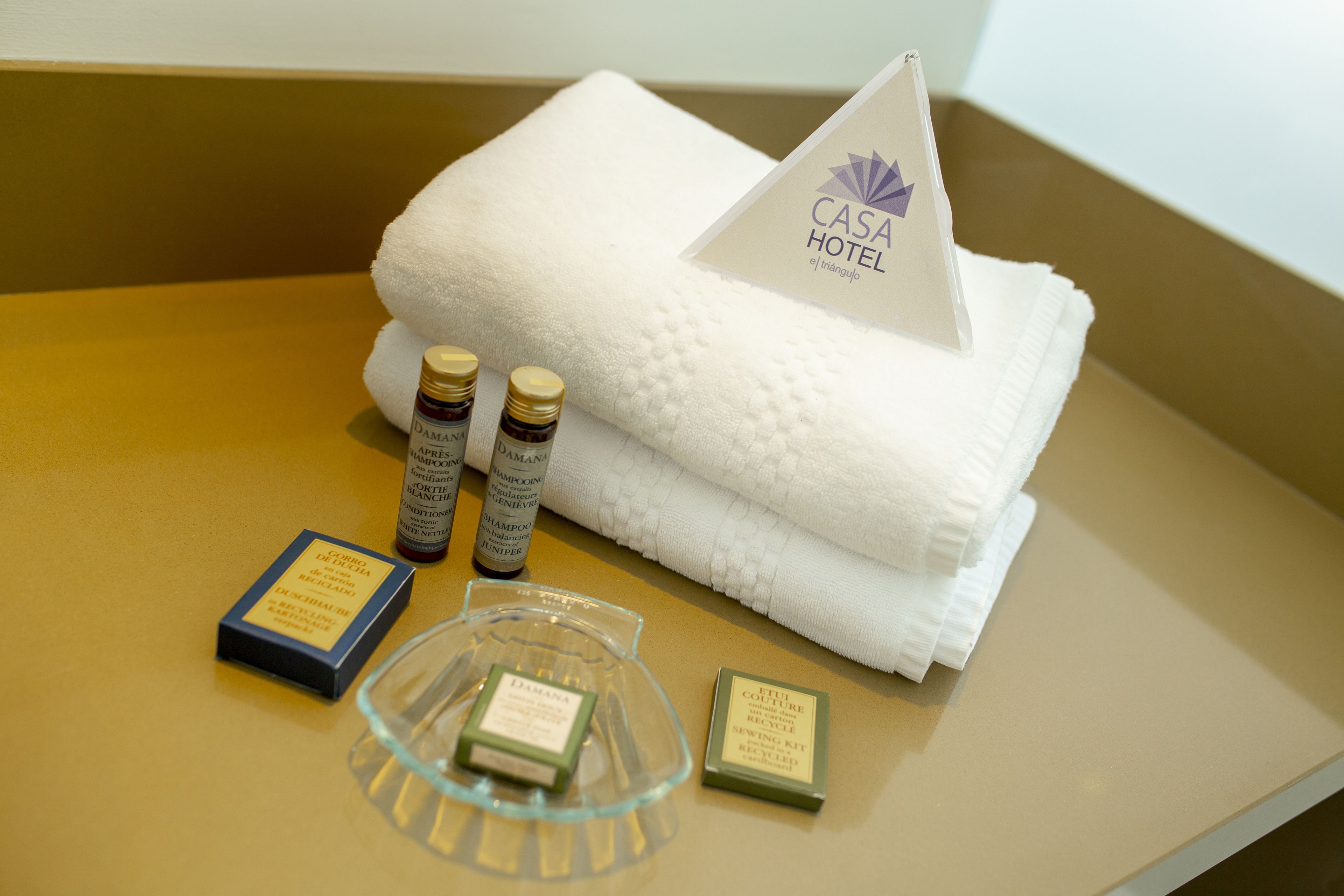 Una toalla blanca doblada con una etiqueta que dice "CASA HOTEL", dos botellas de champú y un pequeño kit de costura sobre una superficie color beige.