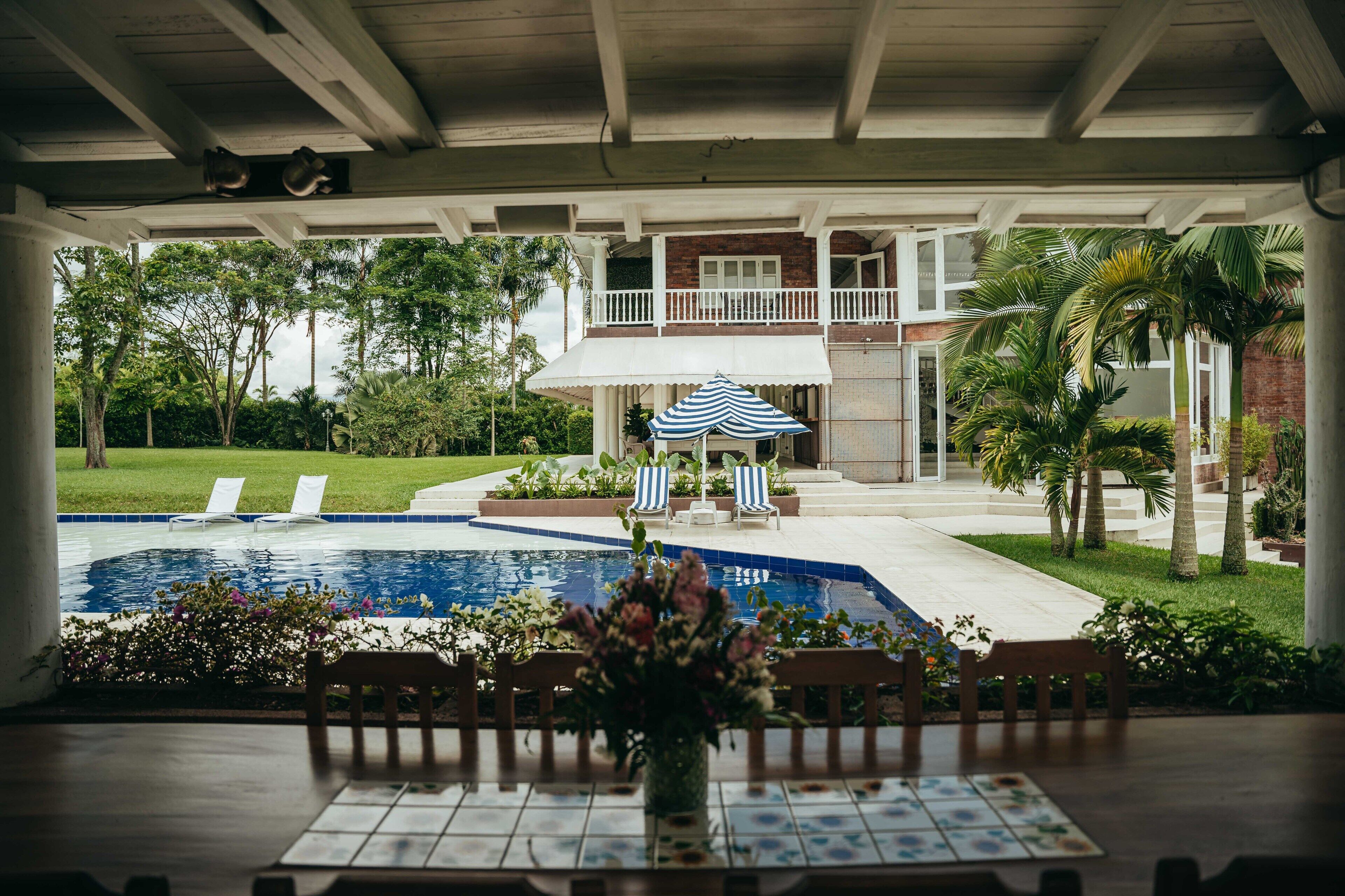 A covered outdoor area with a pool, lounge chairs, and a table with a vase of flowers.