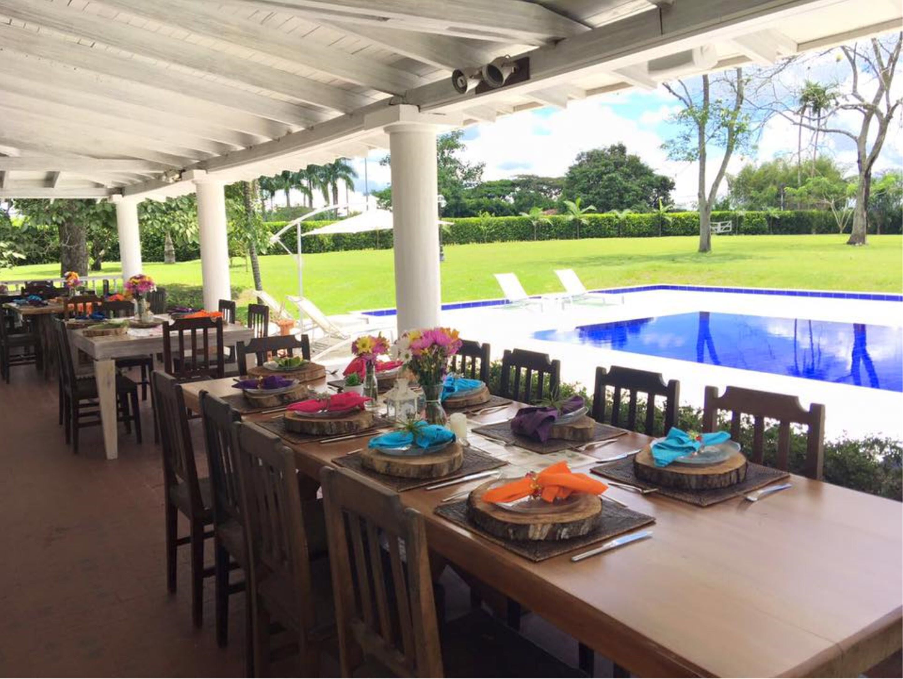 An outdoor dining area with tables set for a meal, overlooking a swimming pool and a well-maintained lawn.