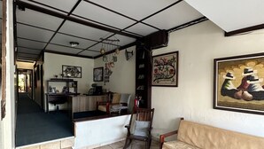 Amplia sala de estar con un sofá, un banco de madera y una silla. Hay un escritorio con silla y una estantería. El piso es de baldosas y el techo tiene un diseño de cuadrícula.