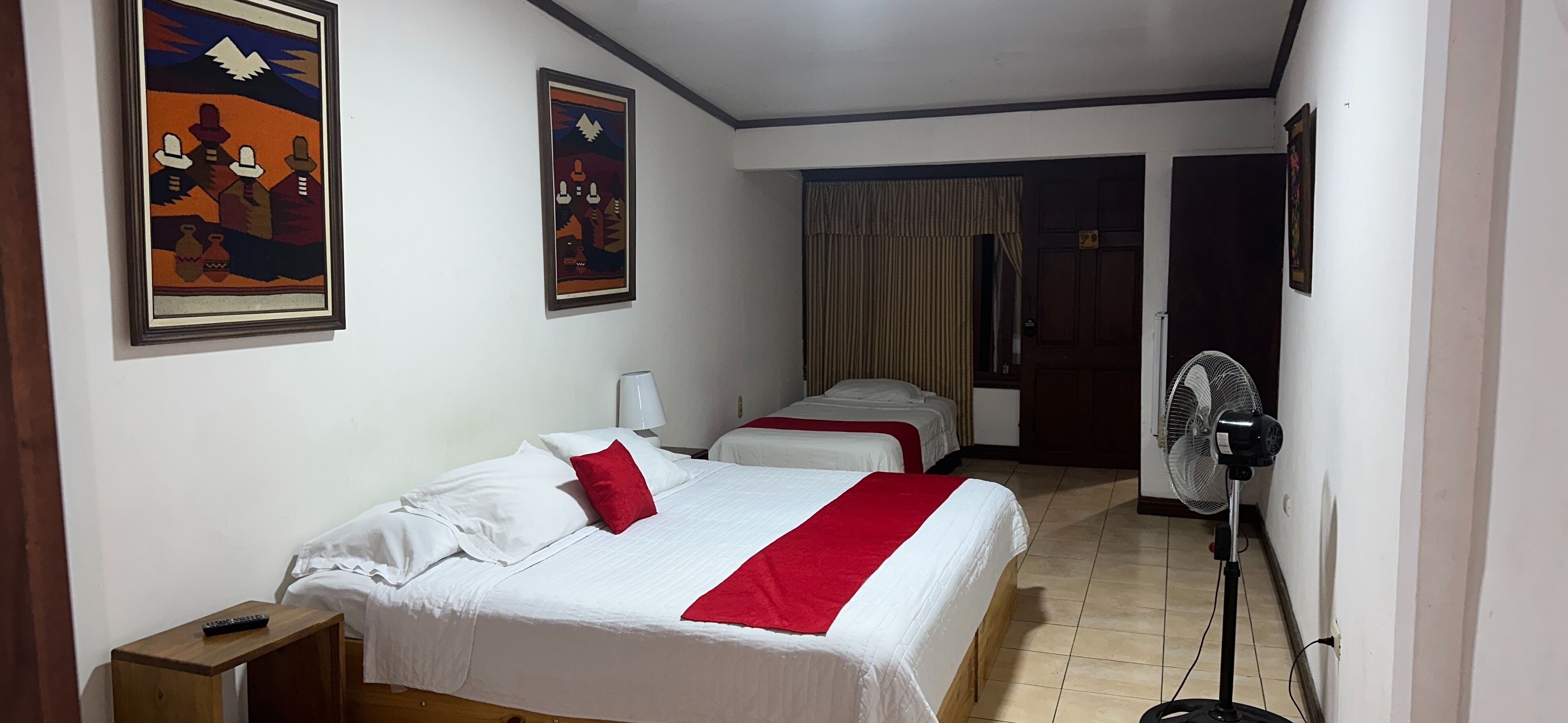 Habitación de hotel con dos camas, un ventilador de pie y dos cuadros en la pared.