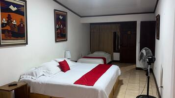 Habitación de hotel con dos camas, un ventilador de pie y dos cuadros en la pared.
