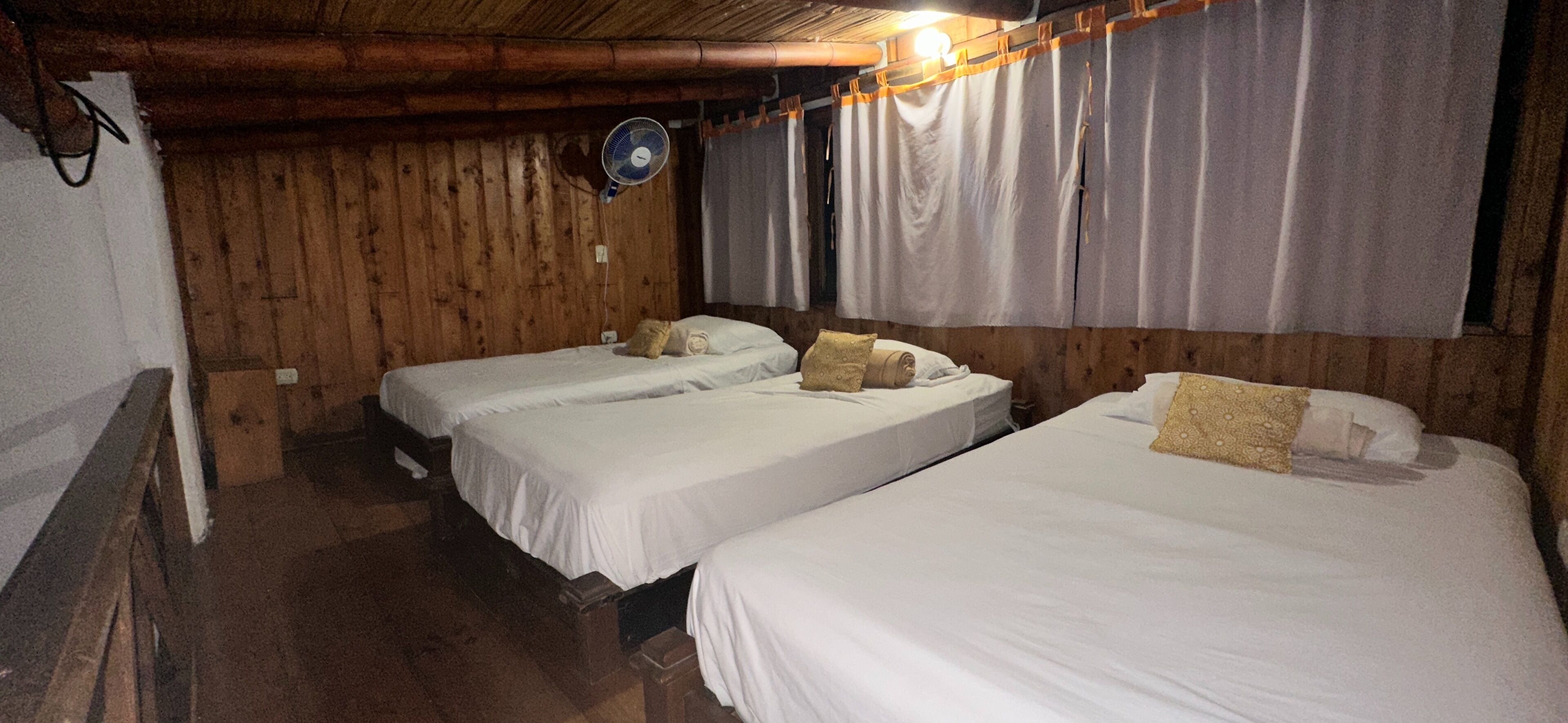 Habitación con dos camas individuales, paredes de madera, ventilador de techo y cortinas.