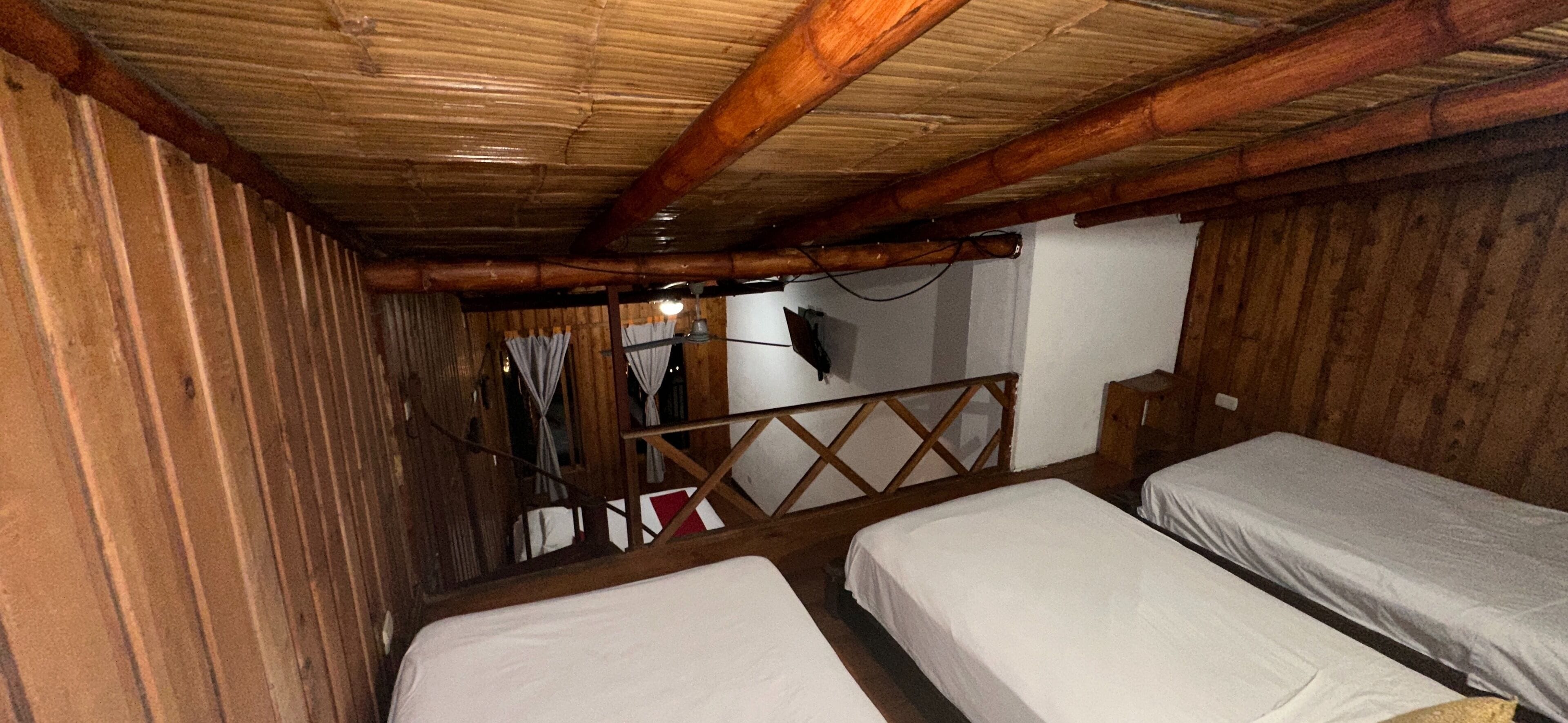 Habitación rústica con paredes de madera y techo inclinado. Hay dos camas individuales con sábanas blancas. Una barandilla de madera separa la zona de dormir de un balcón con vistas.