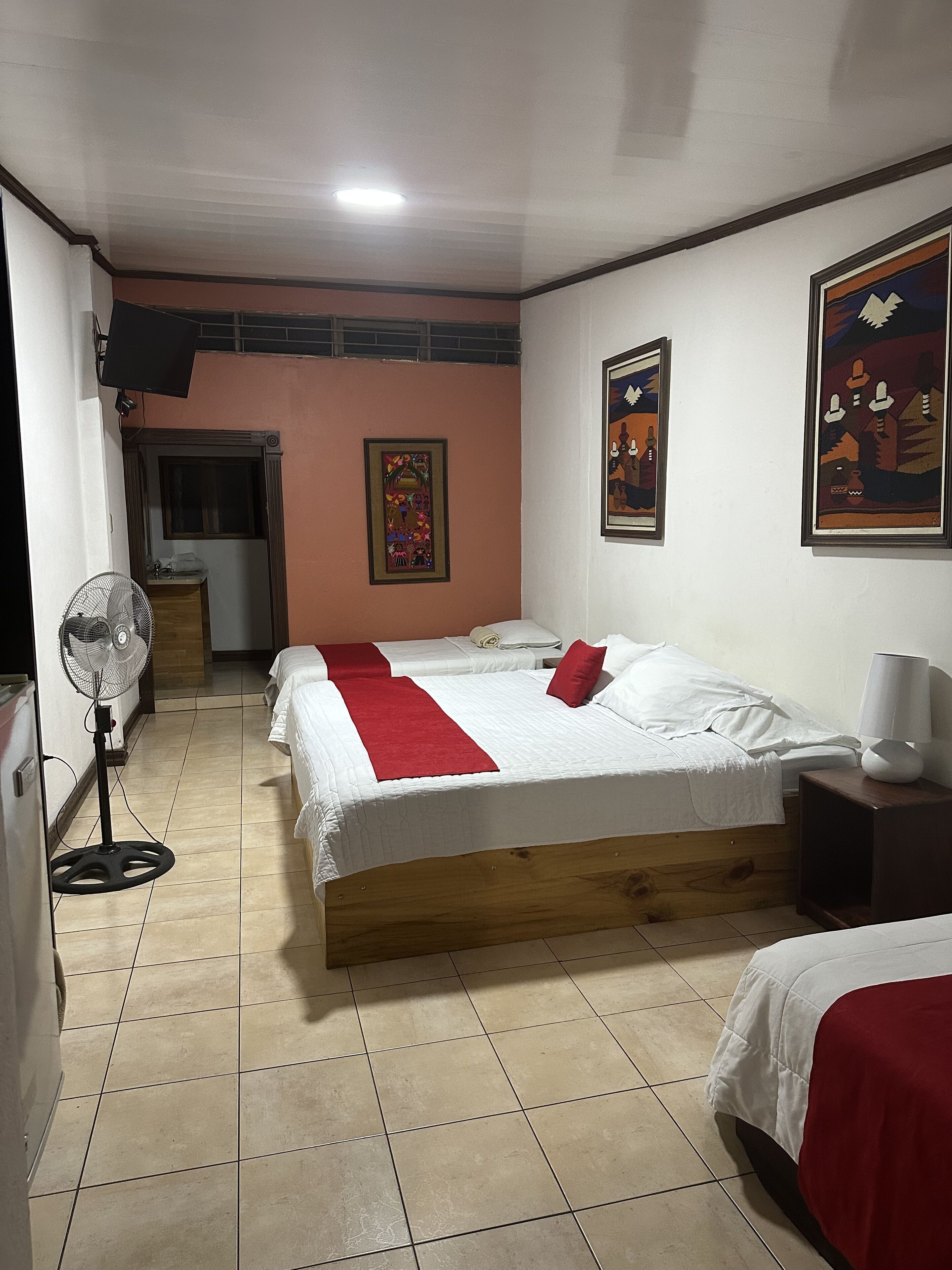 Habitación de hotel con una cama grande, una mesita de noche con lámpara, un ventilador de pie y una ventana con cortinas.