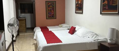 Habitación de hotel con una cama grande, una mesita de noche con lámpara, un ventilador de pie y una ventana con cortinas.