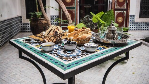 Una mesa servida con pan, frutas y bebidas en un patio con paredes de azulejos y una puerta al fondo.