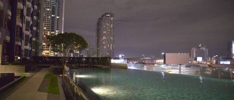 A nighttime view of a modern building complex with a swimming pool, high-rise buildings, and a city skyline in the background.