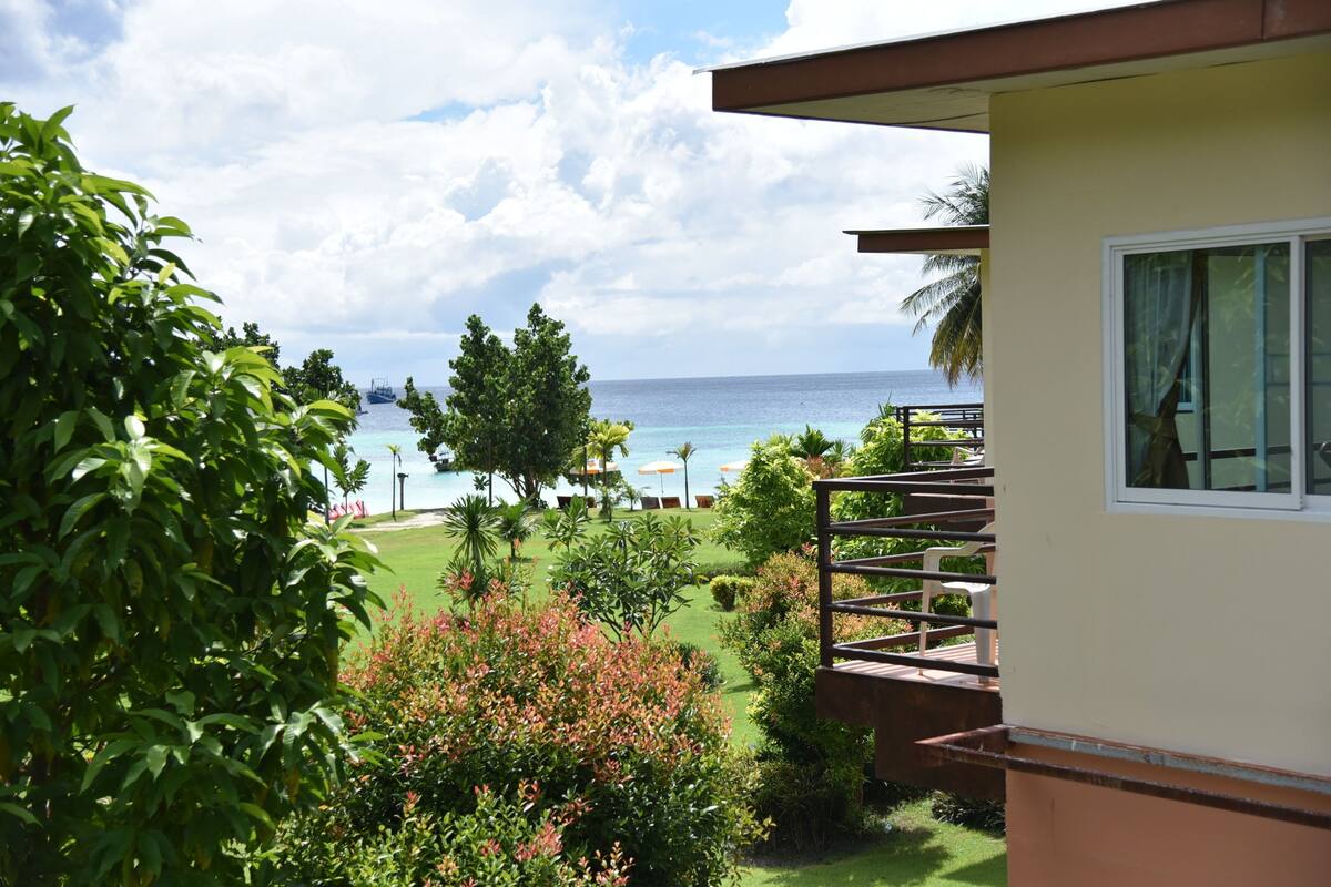 Vista de uma varanda com guarda-corpo, uma pequena área de jardim com diversas plantas, e um vislumbre do oceano e da praia ao fundo.