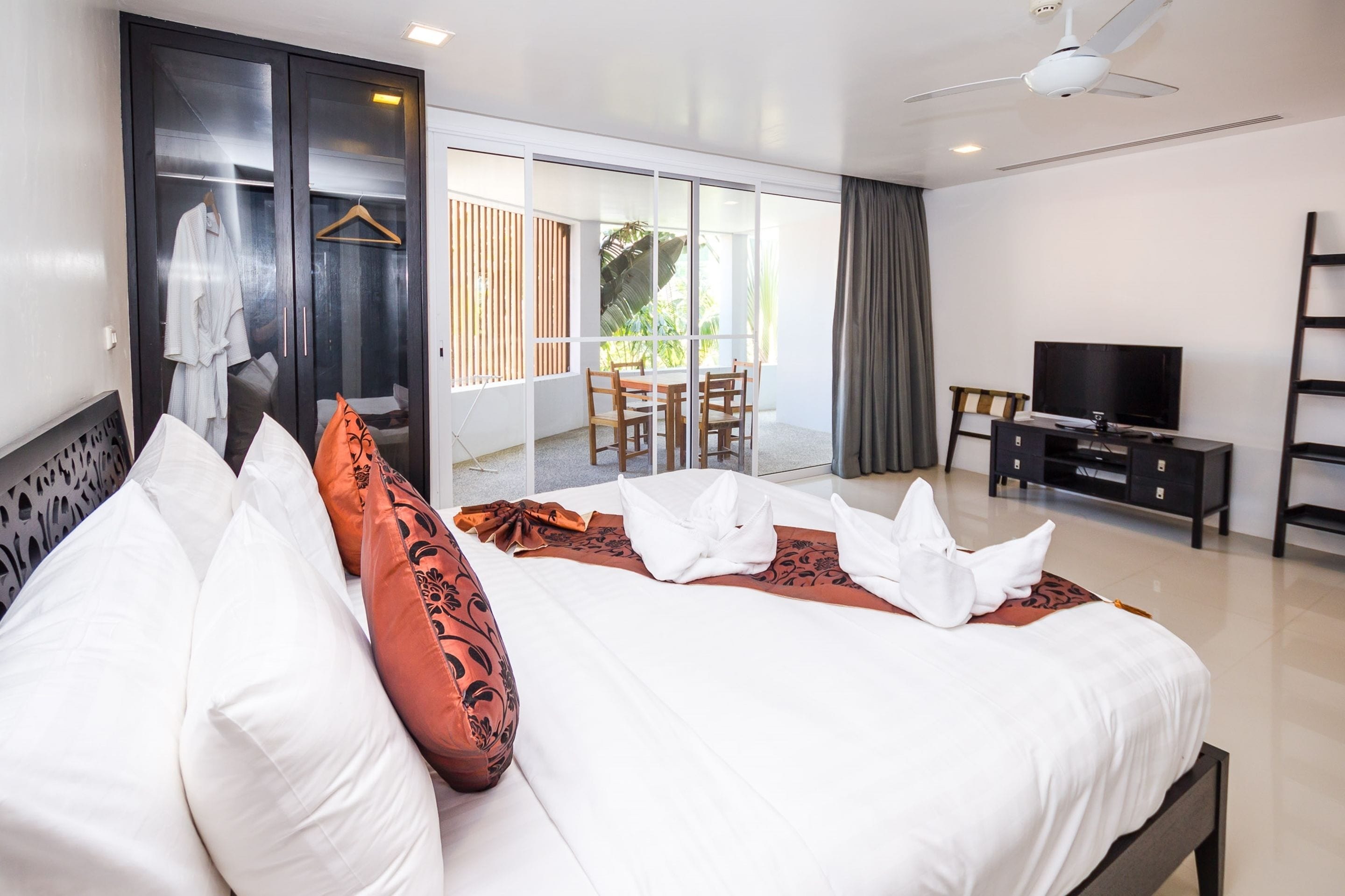 A modern hotel room with a large bed, a TV, a balcony with a table and chairs, and a sliding door leading to the balcony.