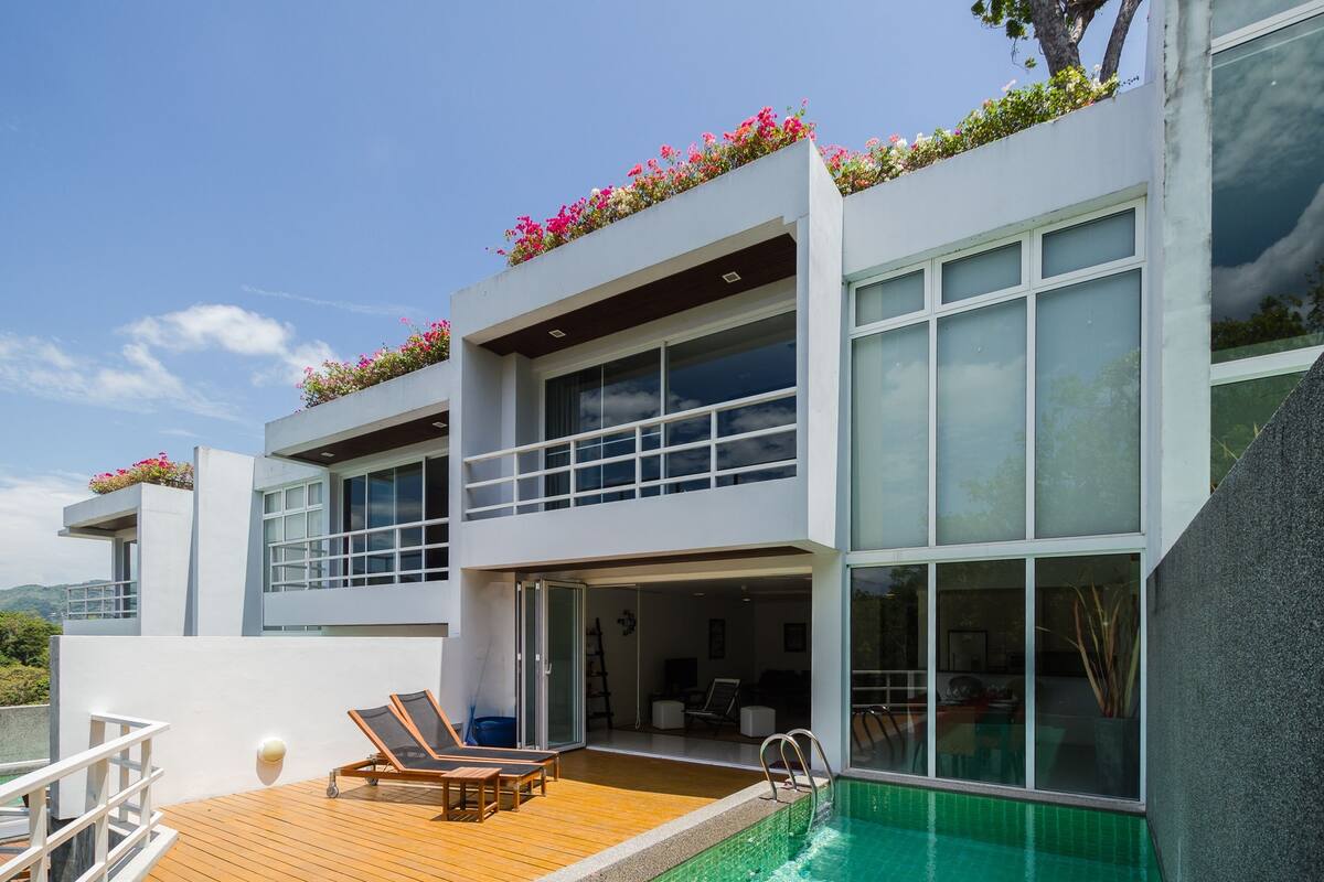A modern two-story building with a rooftop garden, a swimming pool, and a wooden deck with lounge chairs.