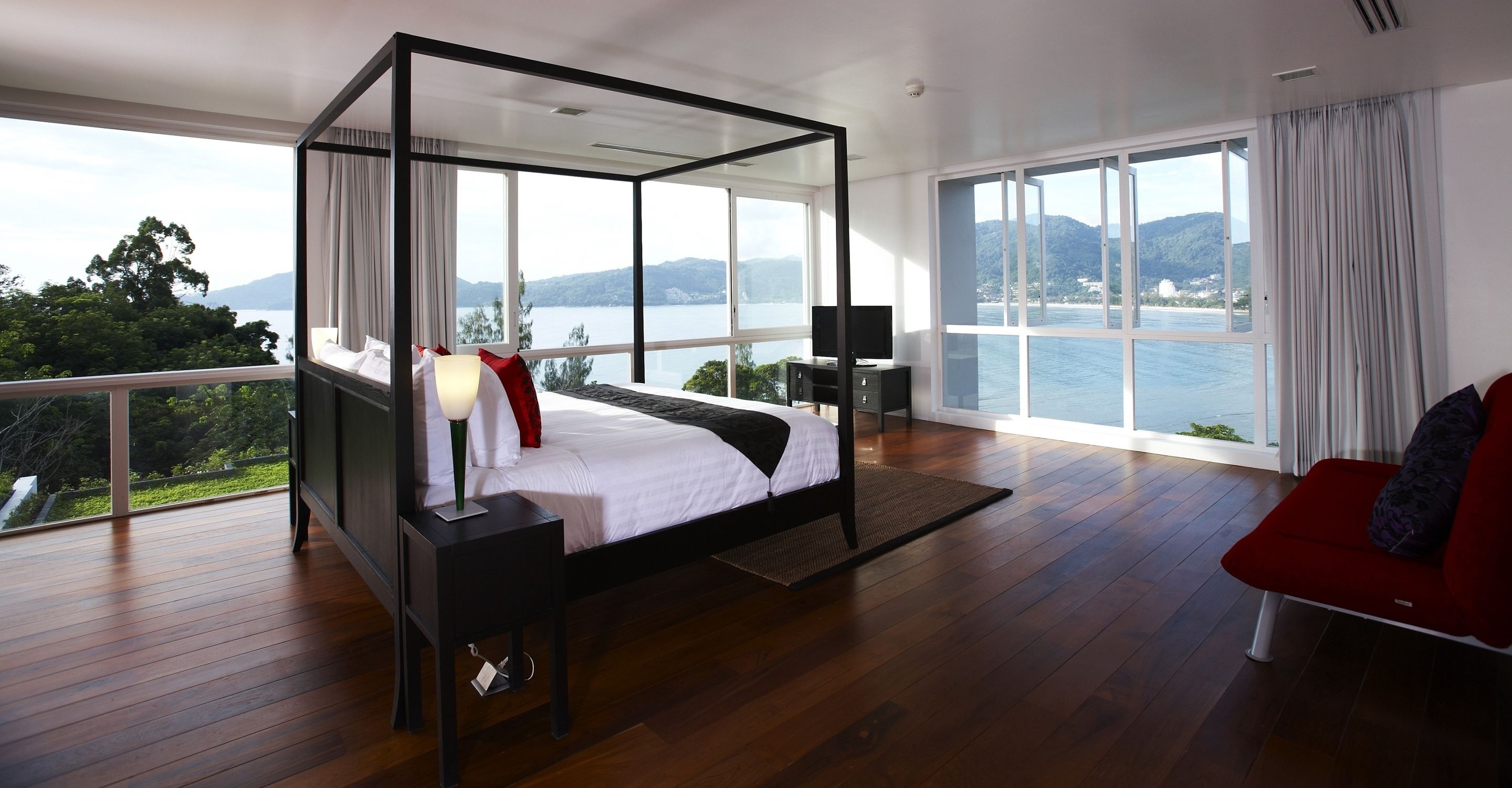A modern bedroom with a four-poster bed, a TV, and a view of the sea.