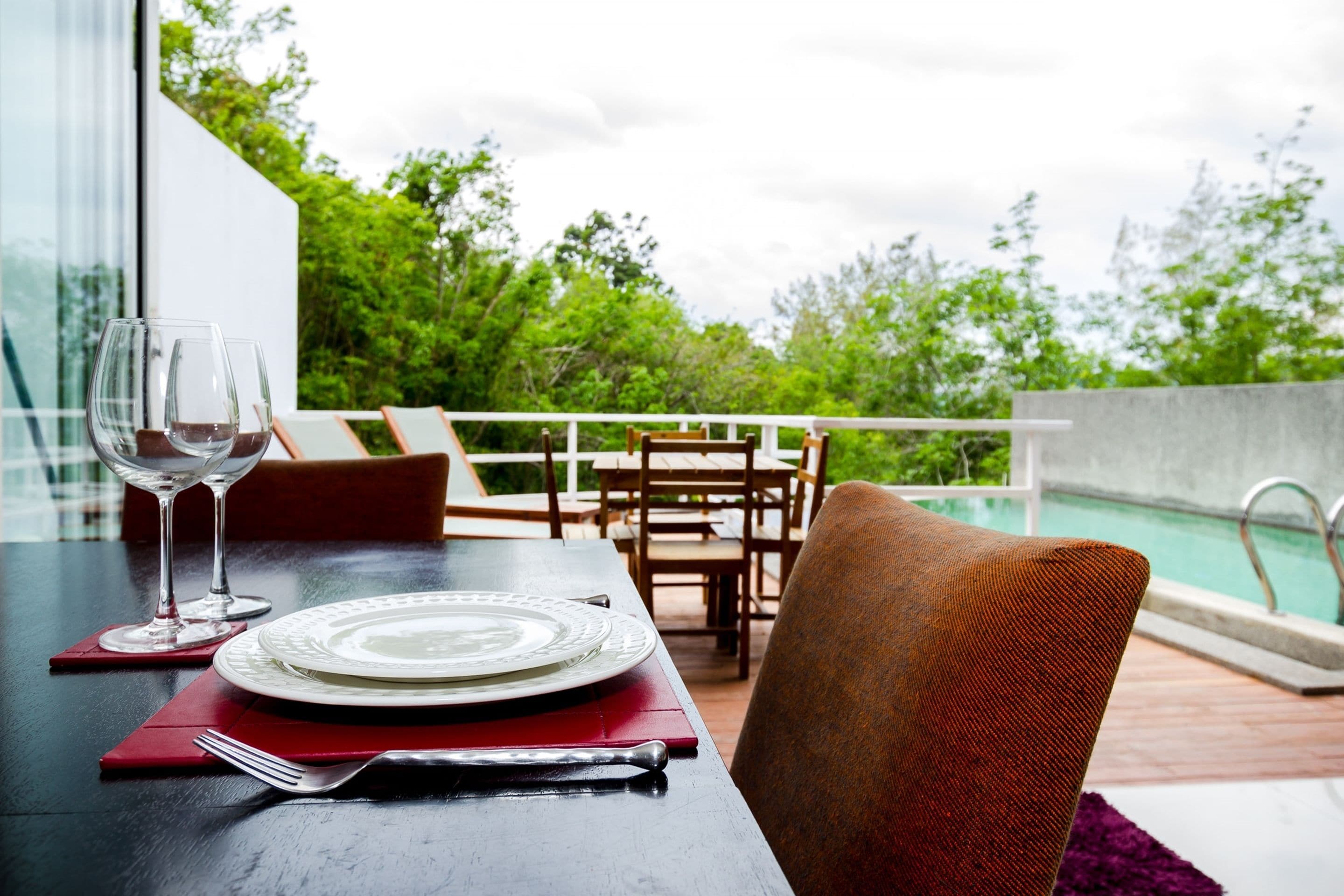 A dining table with a plate, cutlery, and wine glass, overlooking a pool and lush greenery.