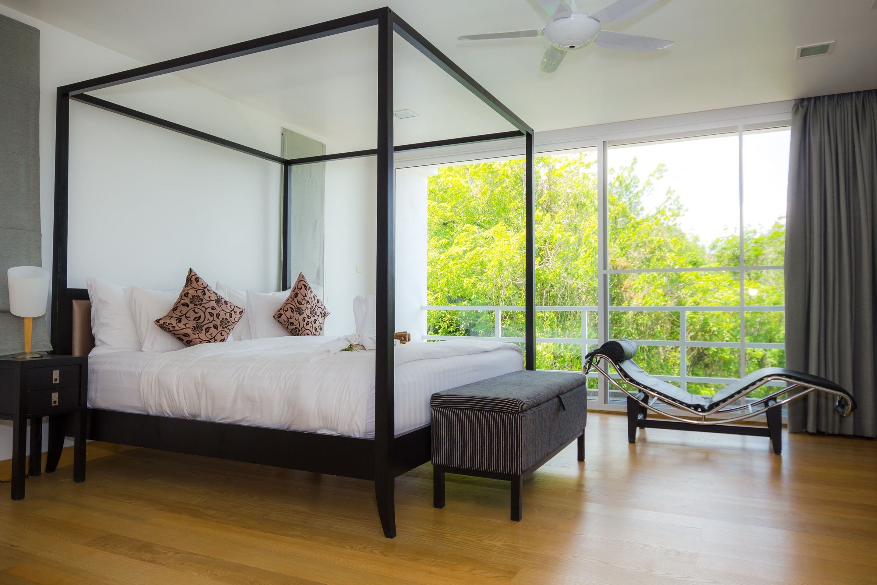 A four-poster bed with a canopy, a bench, and a view of greenery through large windows.