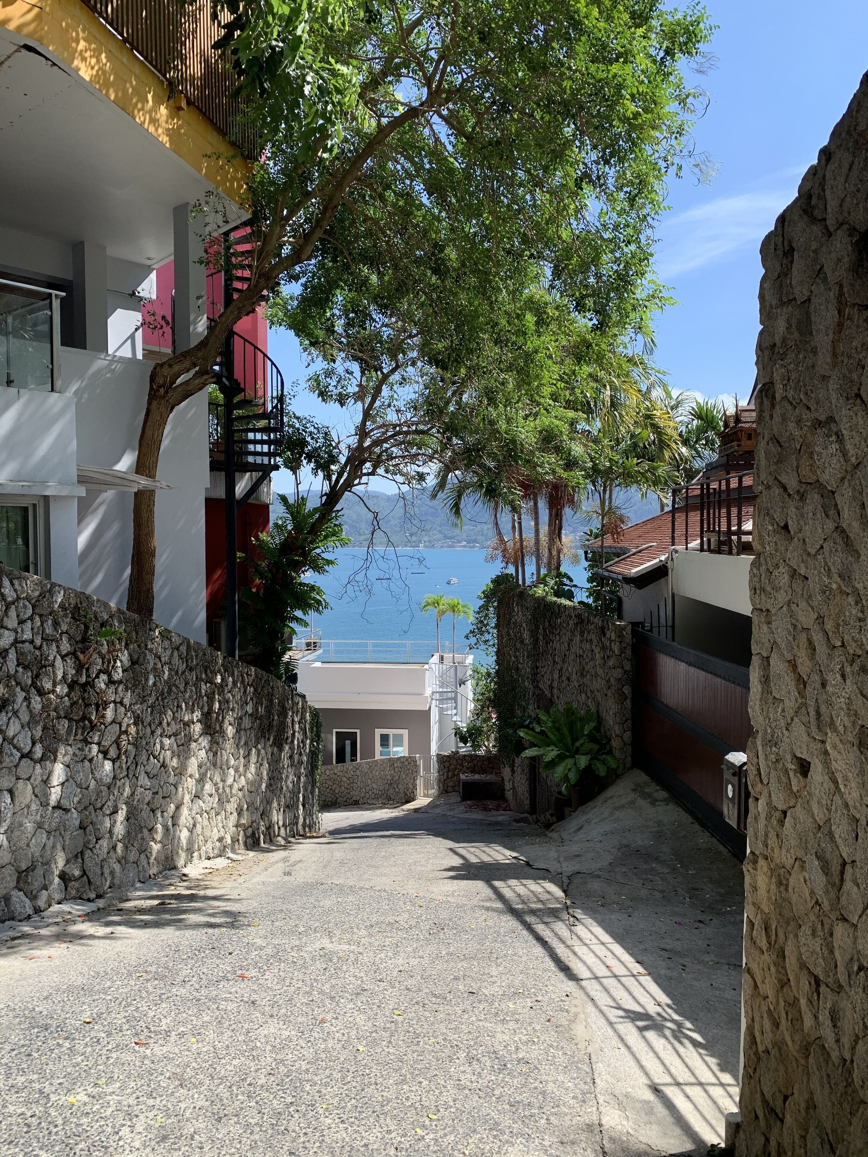A narrow pathway leading to a building with a stone wall on one side and a view of the sea on the other.