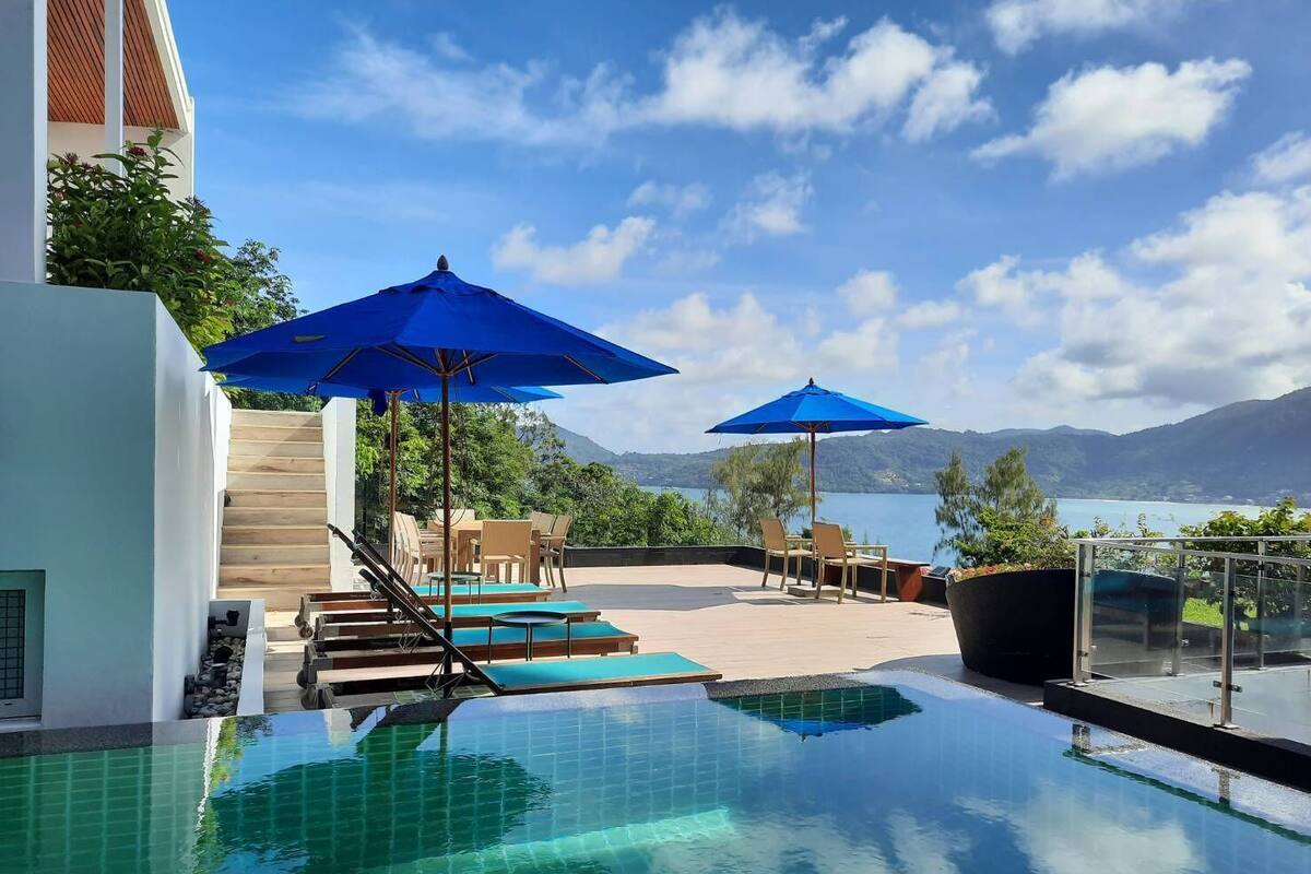 A pool area with blue umbrellas, a view of mountains, and a clear sky.