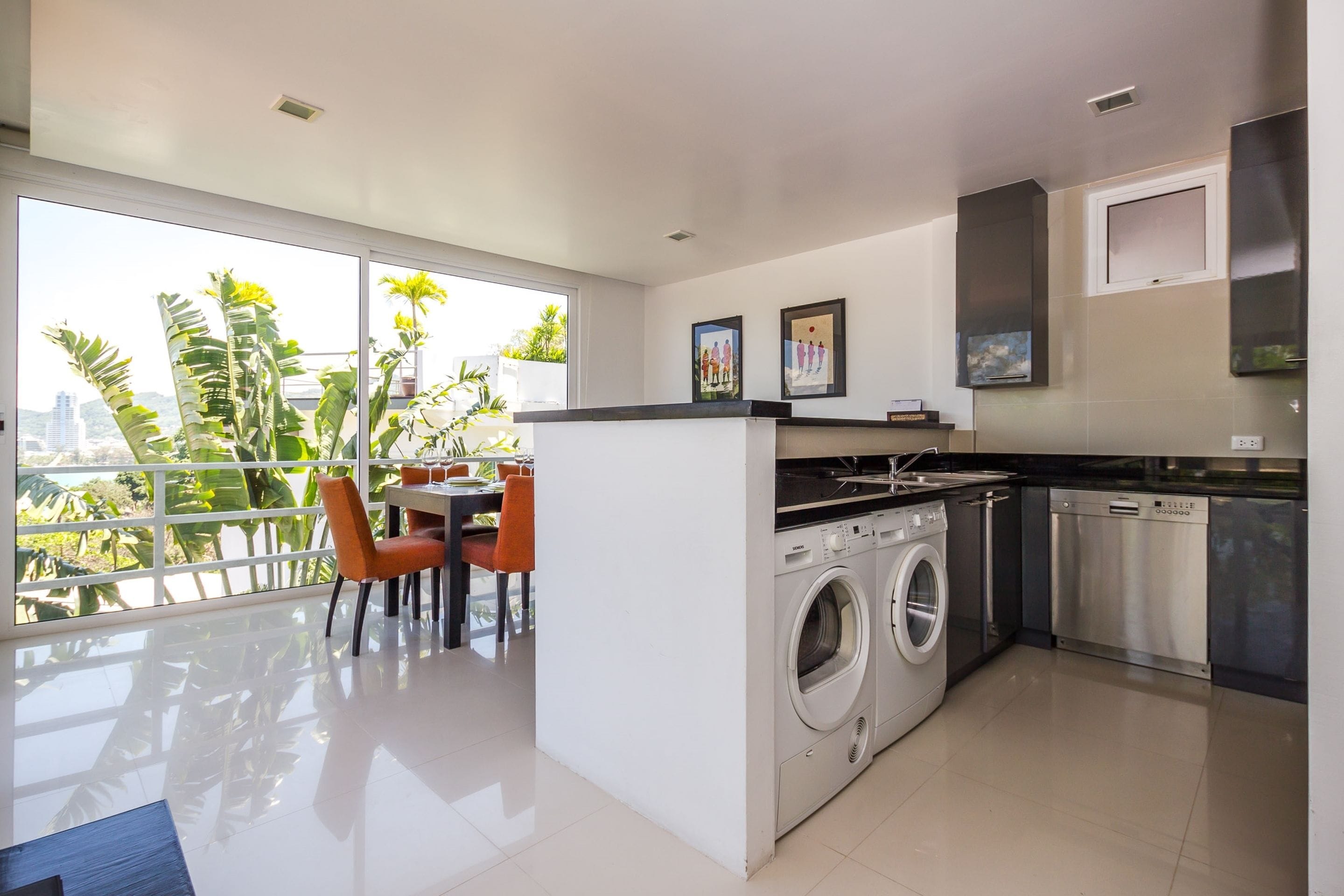 A modern kitchen with a washing machine, a dining area with chairs, and a view of greenery through large windows.