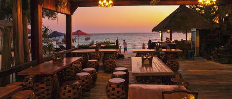 Un área de comedor al aire libre con mesas y taburetes de madera, con vista al mar al atardecer.