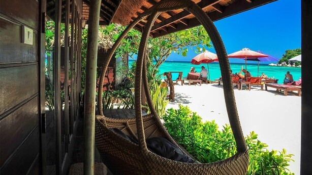 A beachside view with a thatched roof structure, wicker chairs, and a sandy beach with umbrellas. - Anda Resort Koh Lipe (Koh Lipe)