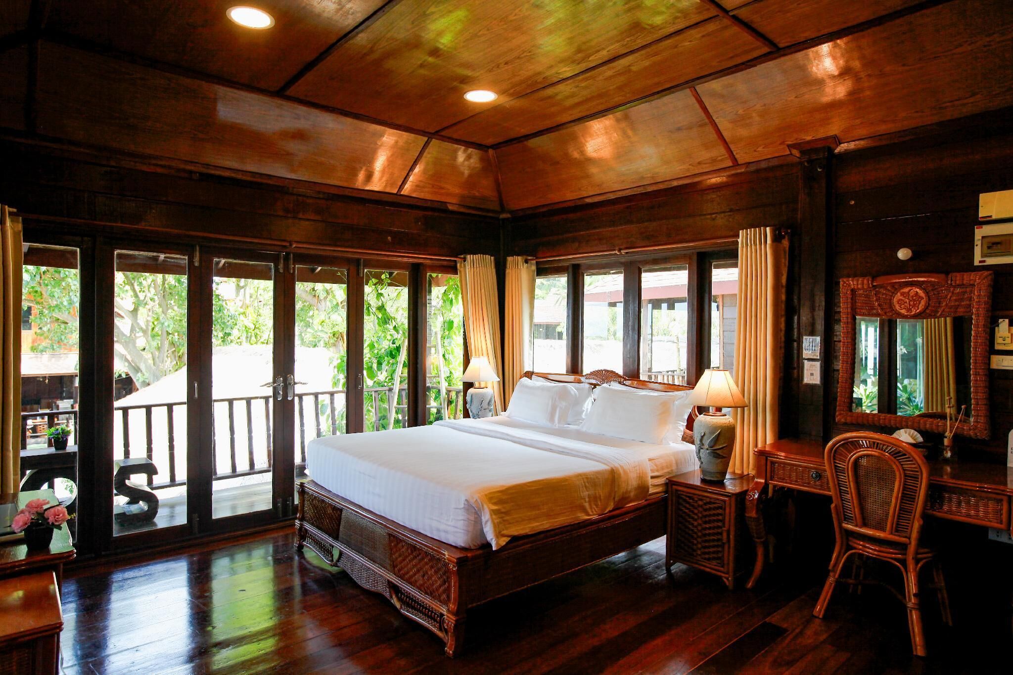 A spacious bedroom with a large bed, a desk, and a chair, featuring wooden flooring and a balcony with a view of greenery.