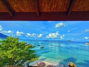 A view from under a wooden structure overlooking a beach with clear turquoise water, boats, and a distant island.