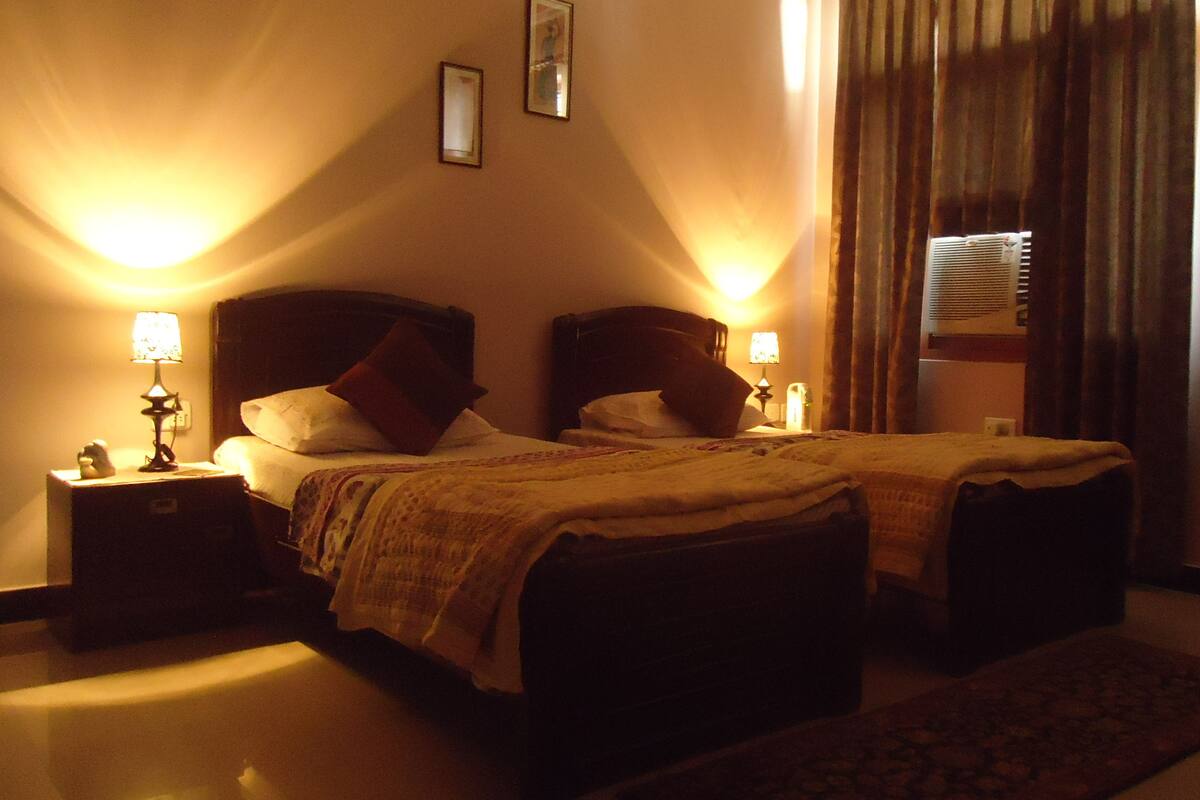 Une chambre d’hôtel avec deux lits, des tables de chevet, des lampes, une fenêtre avec des rideaux et un climatiseur fixé au mur.