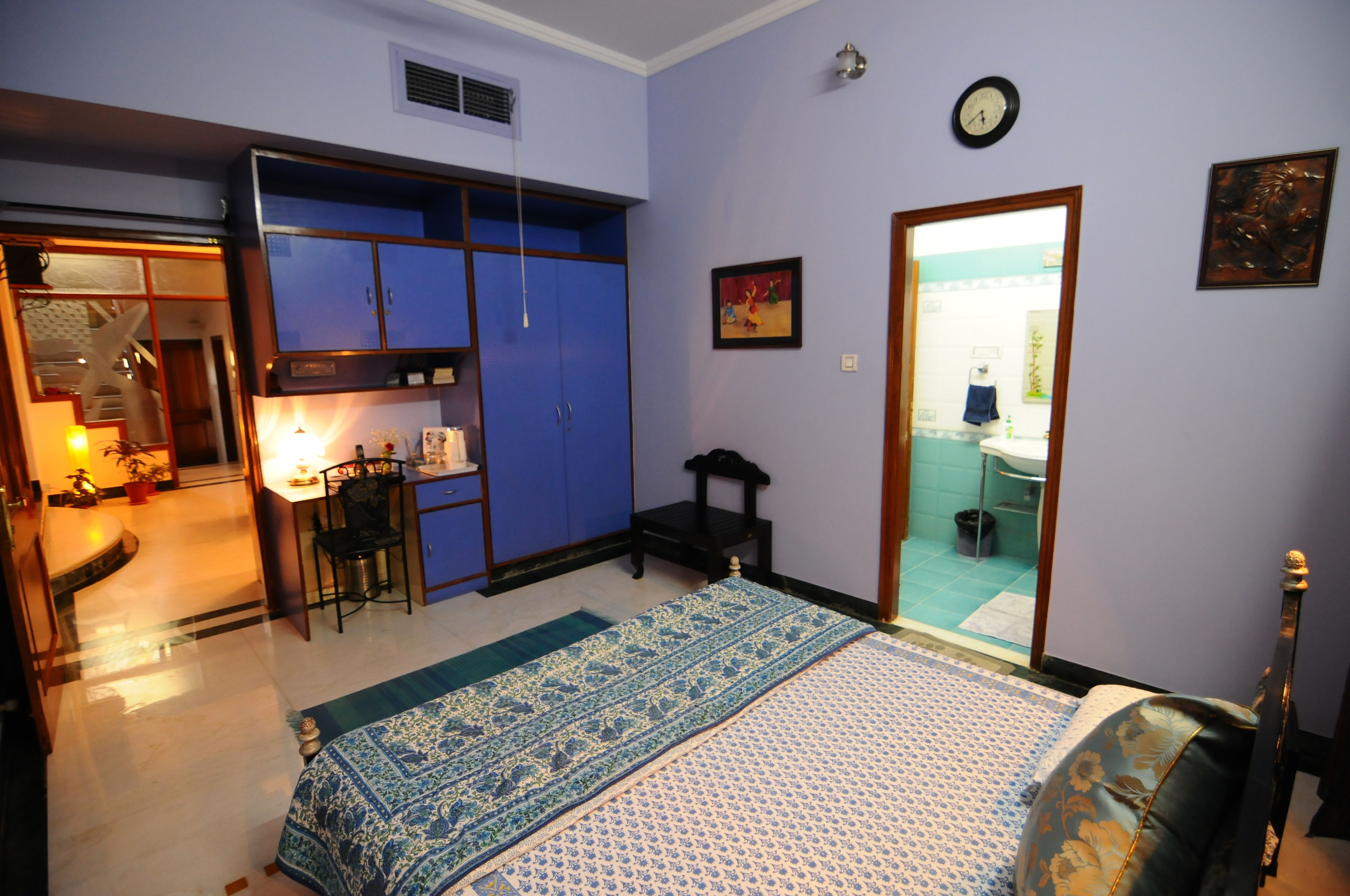A bedroom with a bed, a desk, and a chair. There is a bathroom visible through a glass door.