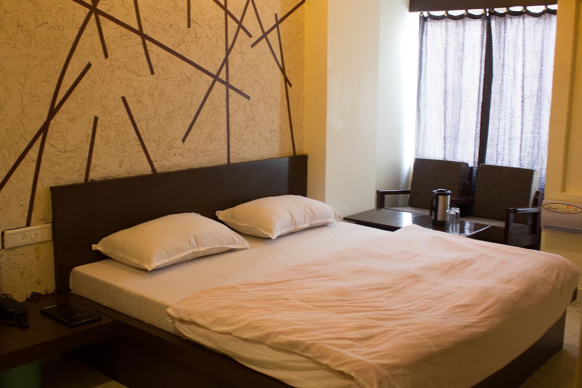 A neatly made bed with white linens, a dark headboard, and two pillows. There is a bedside table with a lamp, a chair, and a small table with a water bottle and a glass. The room has a simple, clean design with a plain wall and a window with curtains.