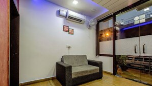 Una habitación con un sofá gris, un equipo de aire acondicionado, un ventilador y una ventana con vista a un edificio.