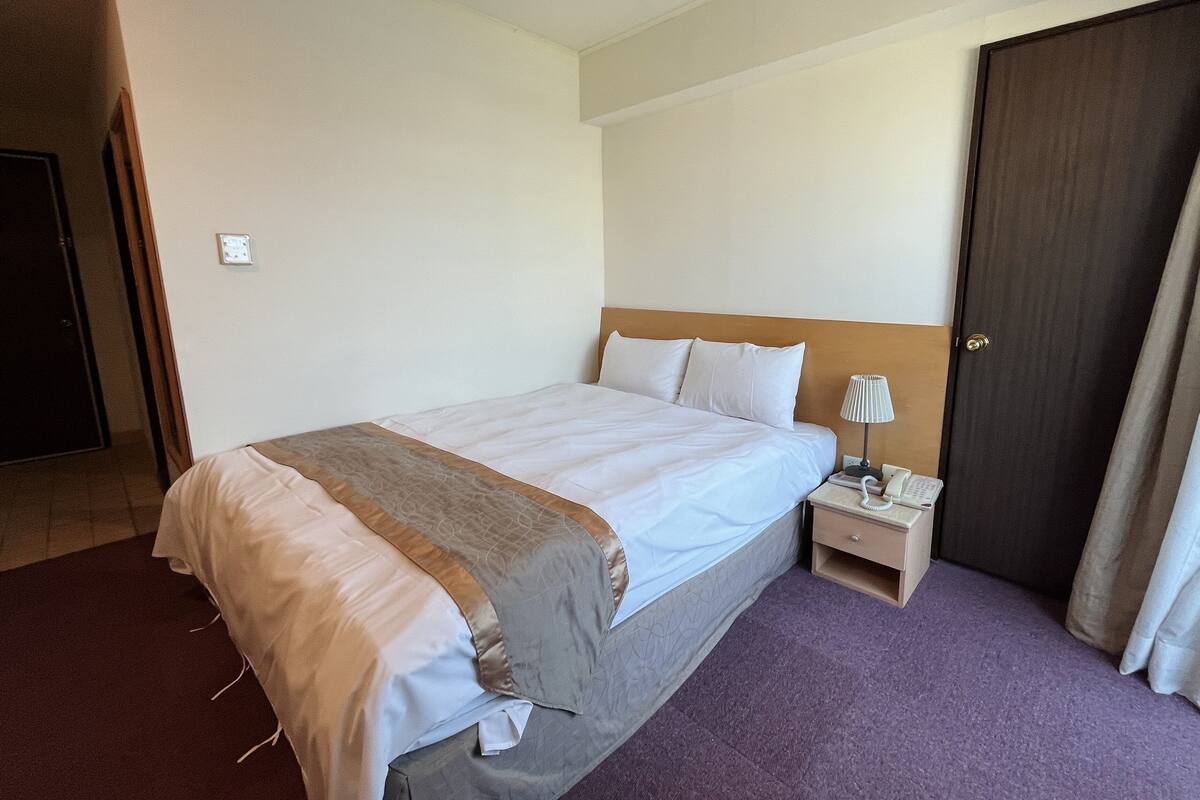 A hotel room with a large bed, a bedside table with a lamp, a phone, and a door.